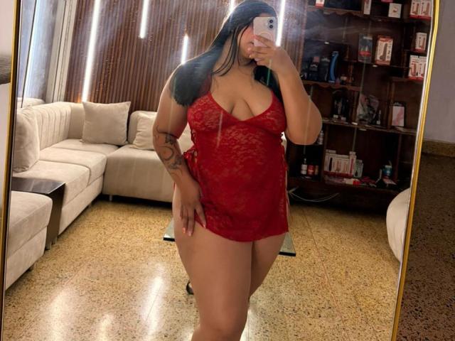 DakotaJhons69 with big tits in a red lace babydoll, taking a mirror selfie, showing a tattoo on her arm, long black hair, and holding a white phone.