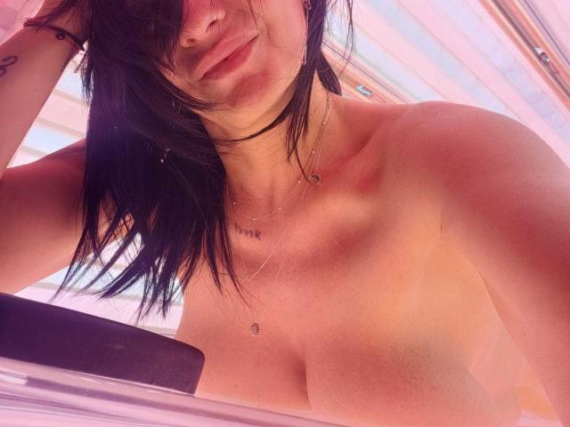 Close-up of IsraKarma, a topless woman with medium-sized breasts, in a tanning bed. Her black hair partially covers her face, and she has a small nose piercing. She wears a black choker necklace and a delicate necklace with a small heart pendant. Her skin has a pinkish tint from the tanning bed. Her right arm, adorned with a small tattoo, rests on the bed's edge. The image has a warm, pinkish glow. The composition is slightly angled, focusing on her upper body and face.