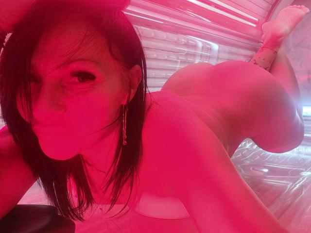 A photo of IsraKarma lying on her stomach in a tanning bed. Her face is slightly turned to the camera, with long black hair partially covering her right eye. She wears gold earrings and a white bikini, exposing her back and buttocks. Her skin is illuminated by pink tanning bed lights, highlighting her toned, curvy physique. Her feet are raised, and she rests on her elbows. The tanning bed's clear plastic walls are visible in the background. The overall composition emphasizes her body and the tanning bed's light.