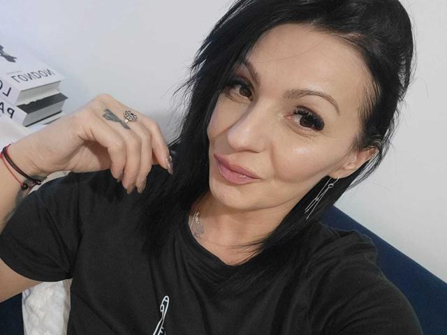 IsraKarma has black hair, brown eyes, and a tattoo on her right ring finger. She's wearing a black shirt, silver earrings, and a subtle smile. Her tits are not visible.