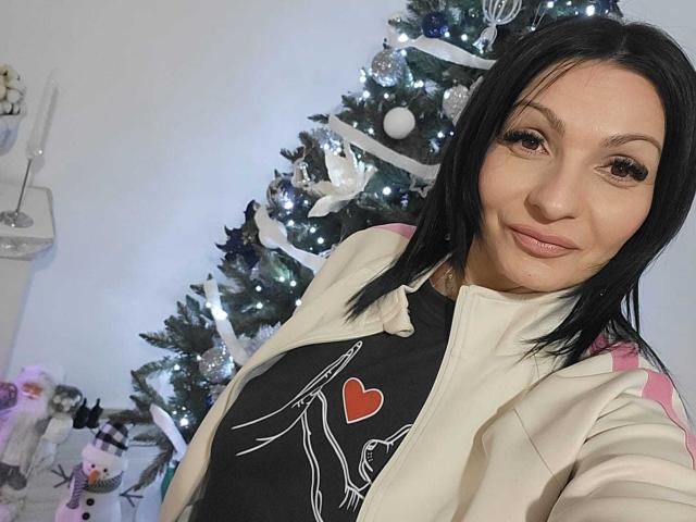 IsraKarma, a woman with straight black hair, wears a beige jacket with pink stripes over a black graphic tee featuring a heart and bird design. She has a subtle smile, light makeup, and is positioned in front of a decorated Christmas tree with white and silver ornaments. The image is a selfie, showing her from the chest up. Her skin is light, and she has a slightly pointed nose. The tree is to the right, slightly blurred.