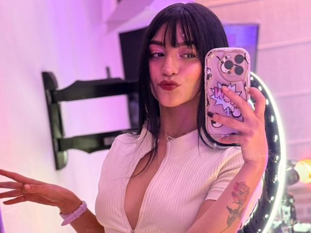 LindaWatsong with black hair, pink makeup, and red lipstick, pouting, wearing a white, ribbed, low-cut top, showing her boobs, holding a phone with a cute case, tattoo on hand.