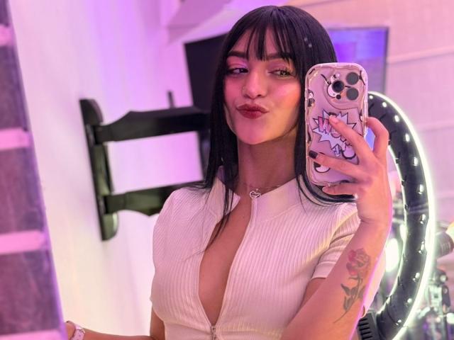 LindaWatsong with black hair, pouty lips, wearing a deep V-neck ribbed top, holding a phone with a cute cartoon skull case, showing a tattoo on her right arm.