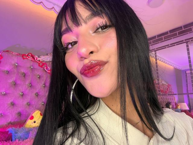 LindaWatsong with glossy pink lips, black straight hair, and hoop earrings, wearing a white top, making a pouty face. Her skin is smooth, and she's in a pink-lit room.
