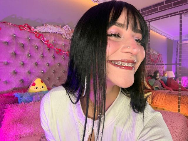 LindaWatsong with black hair, pink eyeliner, nose ring, and metal-grilled teeth, smiling with pink lipstick, wearing a white top, in a pink, purple-lit bedroom.