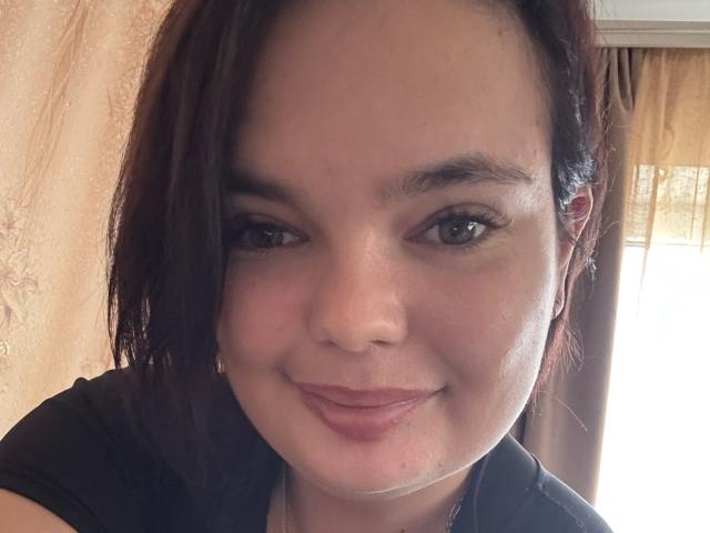 LilaEdna has light brown skin, dark brown eyes, and short brown hair. She's smiling with pink lipstick, wearing a black top. Her small tits are slightly visible.