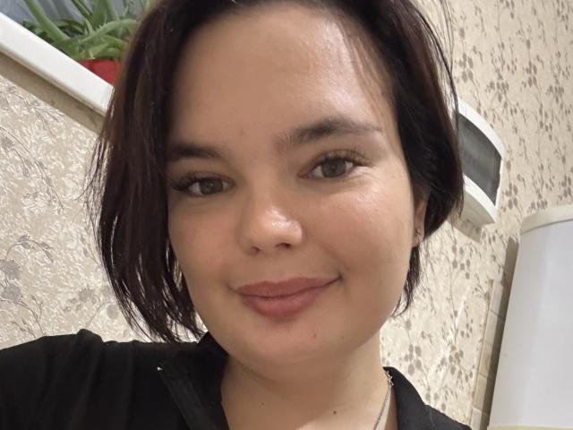 LilaEdna has a cute face with dark brown hair, light skin, and a subtle smile. She's wearing a black top and has small, subtle tits. She looks relaxed.