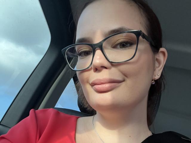 LilaEdna with glasses, pink lipstick, red shirt, black top, small earrings, light skin, brown hair, slight smile, inside car, looking at camera.