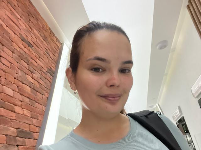LilaEdna with light skin, brown hair in a ponytail, small nose, subtle smile, wearing a light blue sweater, small earrings, black shoulder bag, in a modern indoor setting.