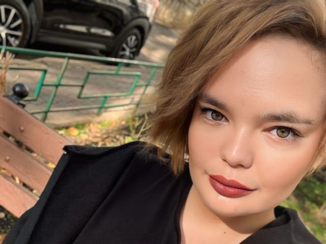 LilaEdna with short brown hair, green eyes, and red lipstick, wearing a black top, has a cute, slightly smug face, and a small beauty mark on her left cheek.