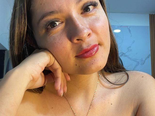 Gabrielafoxxy with shiny red lips, brown eyes, and a small nose piercing, resting her chin on her hand, showing her bare shoulders and a gold necklace.