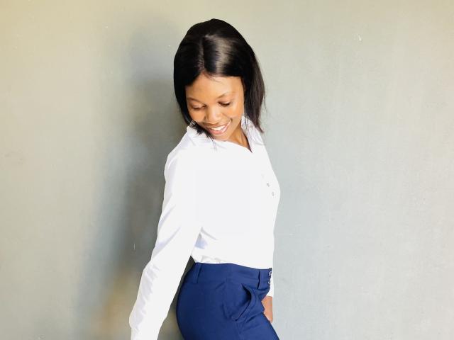 FaithAngel has dark, straight hair, a white shirt, and blue pants, smiling with eyes closed, standing against a plain wall, showing a cute, happy, and confident look.