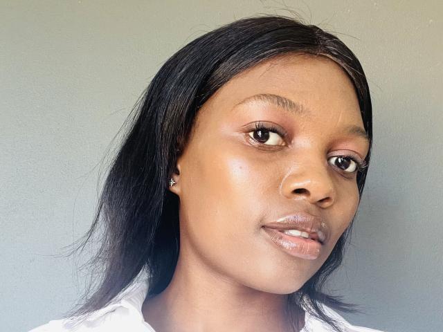 FaithAngel has smooth brown skin, black shoulder-length hair, brown eyes, and shiny pinkish lips. She's wearing a white shirt and has small diamond earrings.