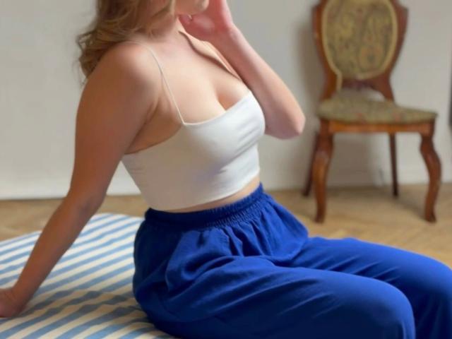AstiAngel-hot with tits in a white crop top, blue pants, sitting on a striped mat, hand near face, wooden chair with patterned cushion in the background.