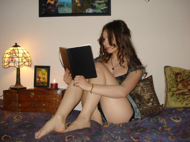 LanaLovely, a young woman with wavy brown hair, sits on a bed reading a black book. She's wearing a dark, short-sleeved top and is barefoot, with her knees drawn to her chest. A patterned pillow and a decorative pillow with a fox design are behind her. A wooden nightstand with a red candle and a lamp with a stained glass shade is to her left. The bed has a colorful, patterned blanket. LanaLovely's skin is fair, and she has a subtle smile. The image has a casual, intimate feel.