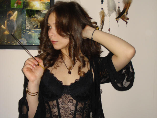 LanaLovely has wavy brown hair and wears a black lace bustier with a sheer black robe. She holds a pencil in her right hand, her left arm raised. Her skin is fair, and she has a necklace with a small dark stone. Feathers are pinned to a wall behind her. The image is a photograph with a slightly blurry effect. Her expression is neutral, and she looks to the side. The overall composition is centered on LanaLovely.