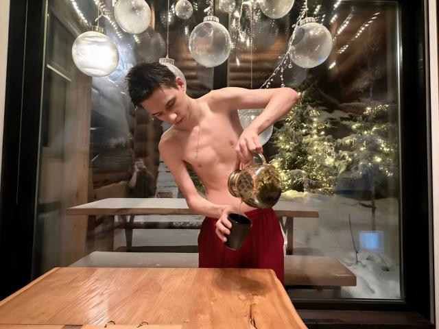 Shirtless Gornits pouring water from a gold trophy into a black cup, wearing red pants, standing in front of a wooden table with hanging glass bulbs.