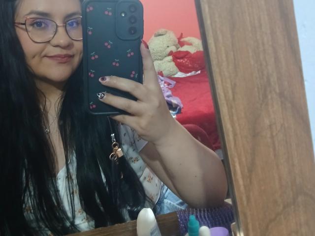 RennattaFox with long black hair, glasses, and a smile, holding a phone with pink flower case, taking a mirror selfie, showing her tits in a white floral top.