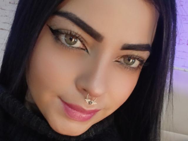 LilithAmoth has green eyes, thick black eyebrows, long black hair, pink lipstick, a nose ring with a small design, and is wearing dark eyeliner.