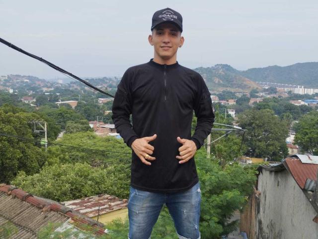 AronRieff stands wearing a black long-sleeve shirt and blue jeans, hands on his belly, with a black cap, on a rooftop, under a cloudy sky.