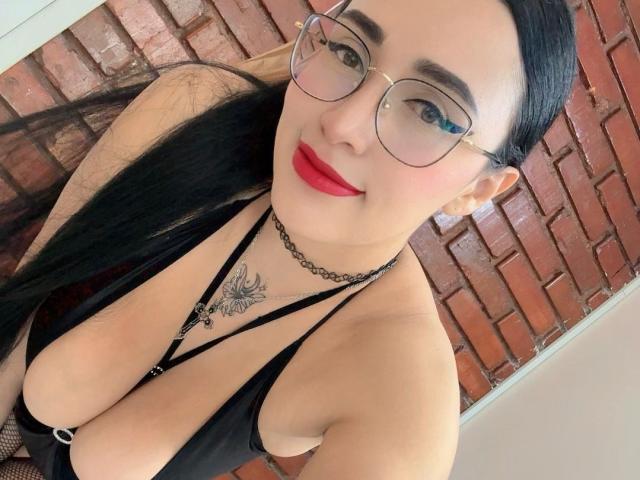 ScarlethSwann with big tits, black strappy top, red lipstick, glasses, black choker, tattoo on chest, long black hair, smiling, brick wall in background, selfie.