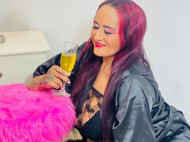 NikolUnlimit with pink and black hair, wearing a black lace top and satin robe, holding a champagne glass, sitting next to a pink furry pillow, smiling.