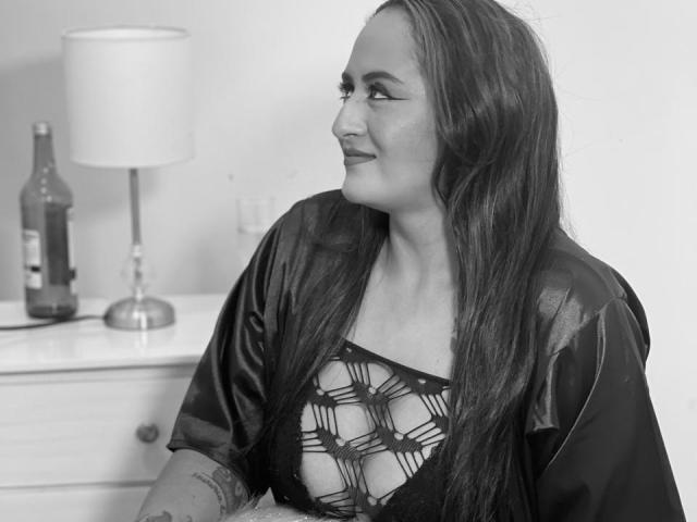 NikolUnlimit, black-and-white photo, with long dark hair, wearing a revealing black lace top, sitting, smiling, bottle and lamp on white dresser in background.