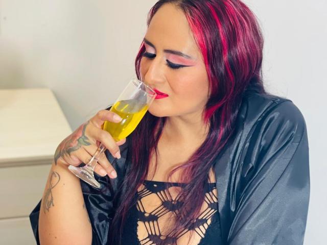 NikolUnlimit with pink and black hair, drinking from a glass of yellow liquid, wearing a black lace top and satin robe, showing tattoos on her hand.