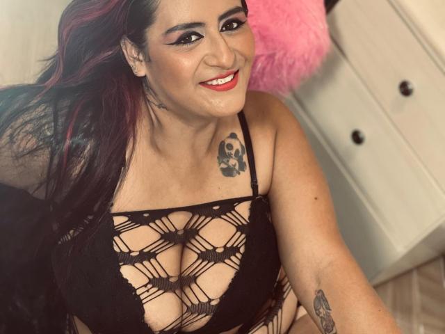 NikolUnlimit with red and black hair, smiling, wearing a black mesh bralette that exposes her tits, skull tattoo on chest, pink fluffy pillow in background.