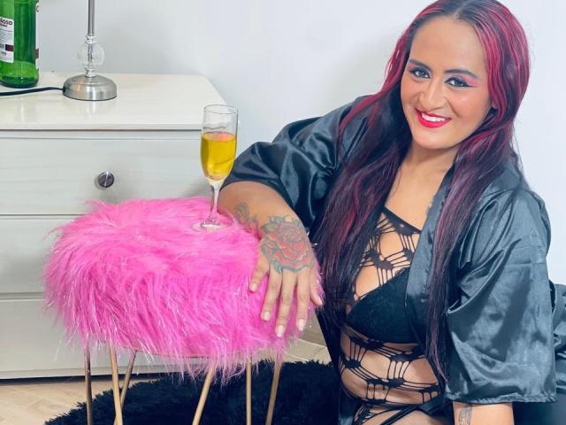 NikolUnlimit with red hair, black lace bra, and shiny robe, leaning on pink furry stool, tattooed hand, smiling, holding champagne glass, showing her tits and ass.