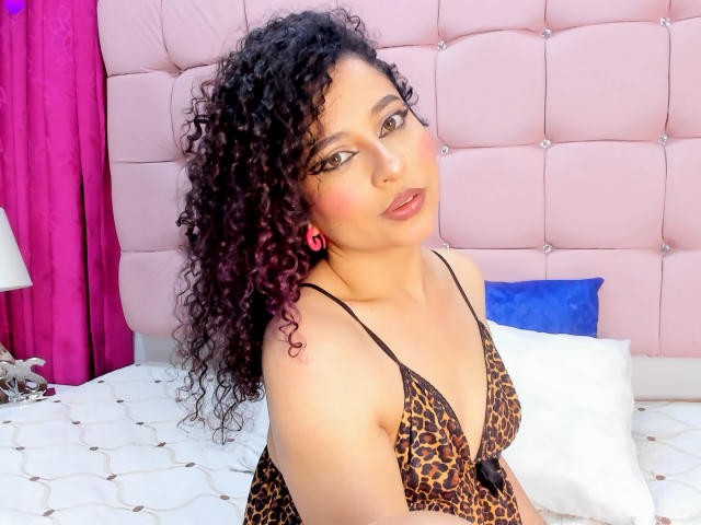 Curly black-haired IrinaSweett with pink lips, wearing a leopard print bra, sitting on a bed, pink headboard, purple curtains, white pillows, and a blue pillow.