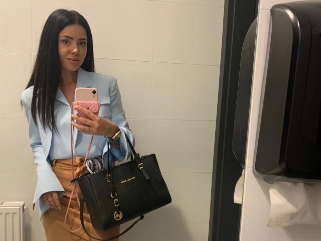NicoleKatze-hot with long black hair, light blue blouse, tan high-waisted skirt, black handbag, pink phone case, holding phone, mirror selfie, smiling, white tiled wall, hand dryer.