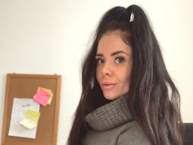 NicoleKatze-hot with long dark hair in pigtails, green eyes, and a gray turtleneck, looking over her shoulder with a slight smile, in a bright office.
