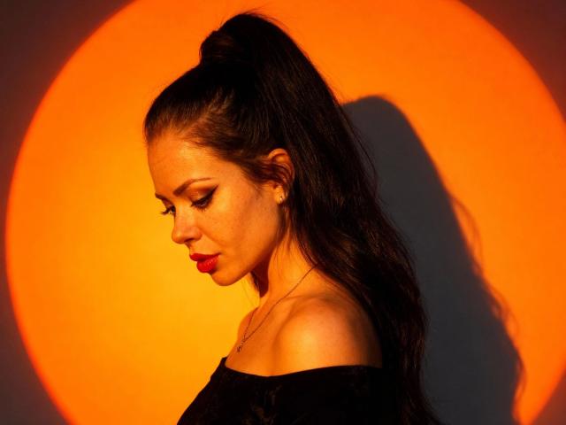 NicoleKatze-hot with dark ponytail, wearing black off-shoulder top, looking down, orange circular light behind, shadow on right, red lipstick, small gold earrings, soft light on skin.