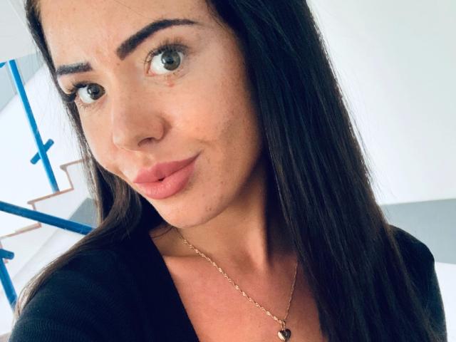 NicoleKatze-hot has long dark hair, green eyes, and pink lips, wearing a black top with a gold necklace. Her nose is slightly upturned. Her boobs are not visible.