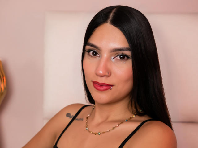 NoemiFerrer with long black hair, red lipstick, and a gold necklace, showing a small tattoo on her right shoulder, wearing a black spaghetti strap top, smiling slightly.