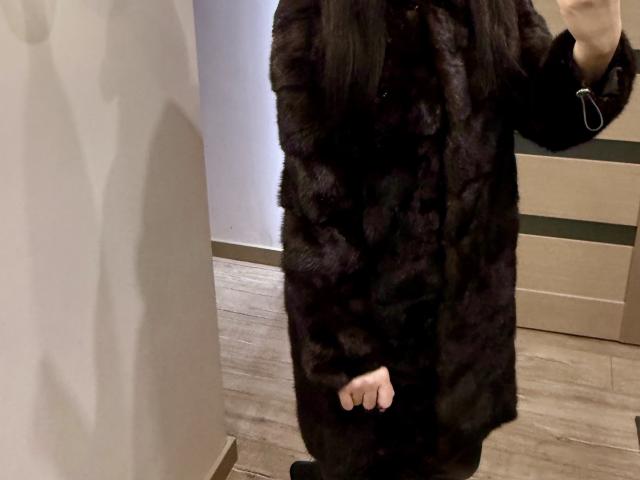 LinaLuchia wearing a dark, fluffy, black fur coat. The coat has a rich texture with visible fur patterns. Her right hand is partially visible at the bottom, with black gloves. The coat covers her torso and extends to mid-thigh. She's standing on wooden floorboards, with a shadow cast on a beige wall to the left. The background includes a wooden cabinet with horizontal slats. The image is cropped to show only her lower torso and coat.