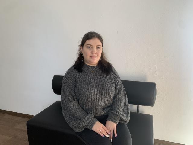 NikaKvitka, with dark brown hair, sits on a black chair wearing a gray knit sweater and black pants, hands resting on her knees, looking straight at the camera.