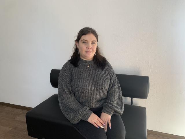 NikaKvitka with dark hair, wearing a gray knit sweater and black pants, sitting on a black chair, hands on lap, wooden floor, white wall, slight smile.