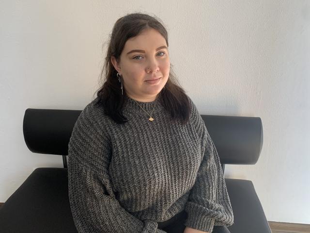 NikaKvitka with dark brown hair, wearing a gray knit sweater, black earrings, and a gold necklace, sitting on a black chair, smiling softly.