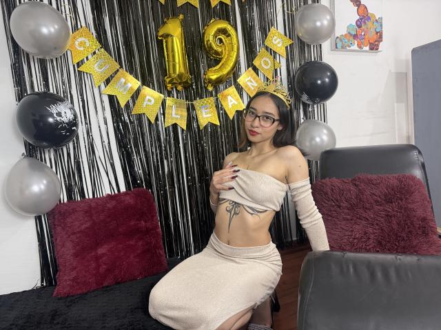 CamiVargas, wearing a beige off-shoulder top and matching skirt, covers her tits with one hand, has a tattoo under her boob, and sits on a black couch.