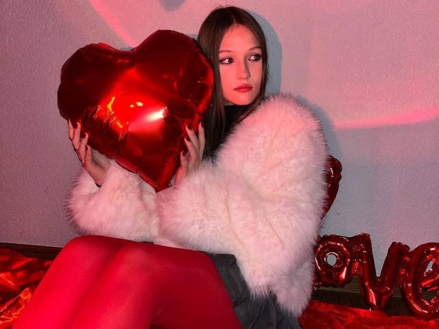Bubochka with red heart balloon, wearing fluffy white jacket, red tights, and grey skirt, sitting on red velvet, looking at camera, with "love" balloon on right.