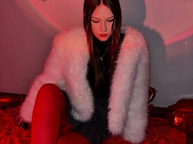 Bubochka sits on a red, textured blanket with a white, fluffy jacket over a black dress. She has long brown hair, red lipstick, and looks down with a neutral expression. Red light casts a shadow on the gray wall behind her. She wears red tights, and a silver necklace is visible. The image has a strong, red-orange color contrast.