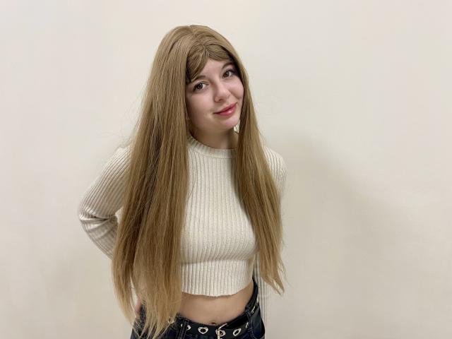 Reginia with long blonde hair, smiling, wearing a white ribbed crop top, hands on hips, black pants with eyelet detail, standing against a plain white wall.