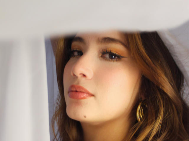 StarDustt's face with light brown wavy hair, orange eyeshadow, black eyeliner, pink lips, small beauty mark under left eye, gold hoop earring, white curtain partially covering face.