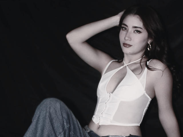 Stardustt with medium tits, wearing a white, button-up, halter crop top and blue jeans, has dark, wavy hair, and is sitting against a black background, looking at camera.