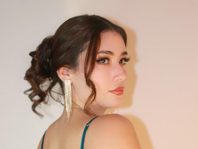 Stardustt has curly brown hair in a ponytail, wearing long, glittering earrings, and looking back with a slight smile, showing her bare shoulders and green strap dress.