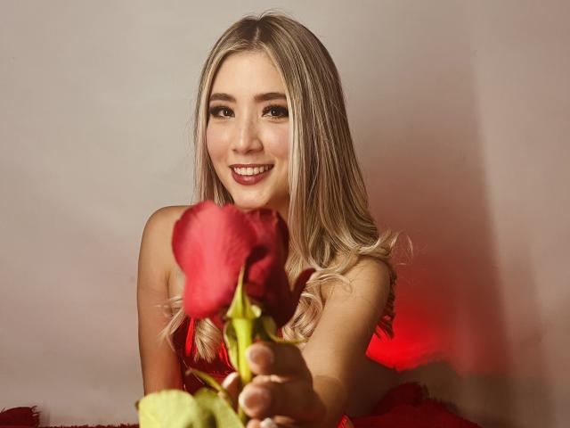 Stardustt with blonde hair, red lipstick, smiling, holding a red rose in front of her, wearing a red dress, soft light, warm colors, slightly blurred background.