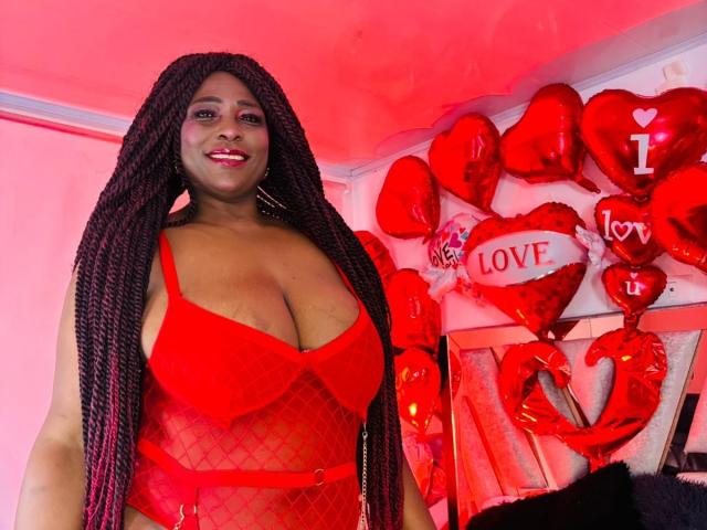 Ebonyy69 with long braids, wearing a red fishnet lingerie top, showcasing her big tits and smiling. Red heart balloons and decorations in the background.