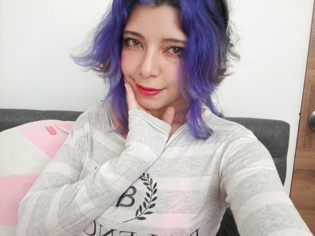 DarckVelvet with purple hair, pink blush, and red lipstick, wearing a gray striped shirt, resting her hand on her cheek, sitting on a gray couch.